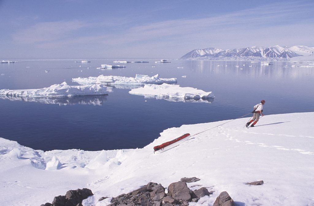 John Dunn sled-hauling at the ice edge on the south coast of Devon Island with a view west to Cape Warrender 1994 – Sandy Briggs photo.