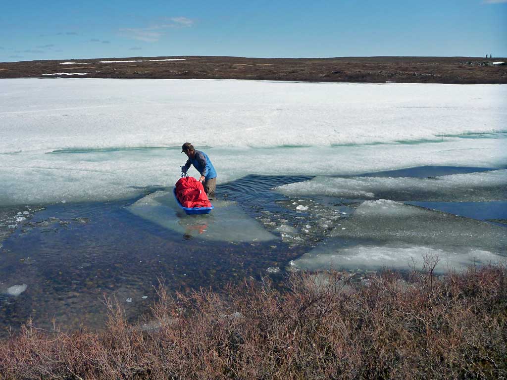 John Dunn uses an ice pan to ferry his sled from the snow-free shore back onto the lake ice. Snowdrift River country in&nbsp;Thaidene Nëné National Park Reserve 2015 – Sandy Briggs photo.